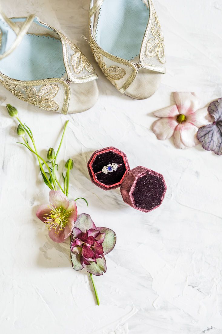 Wedding details with embroidered shoes, flowers, and ring box on white surface.