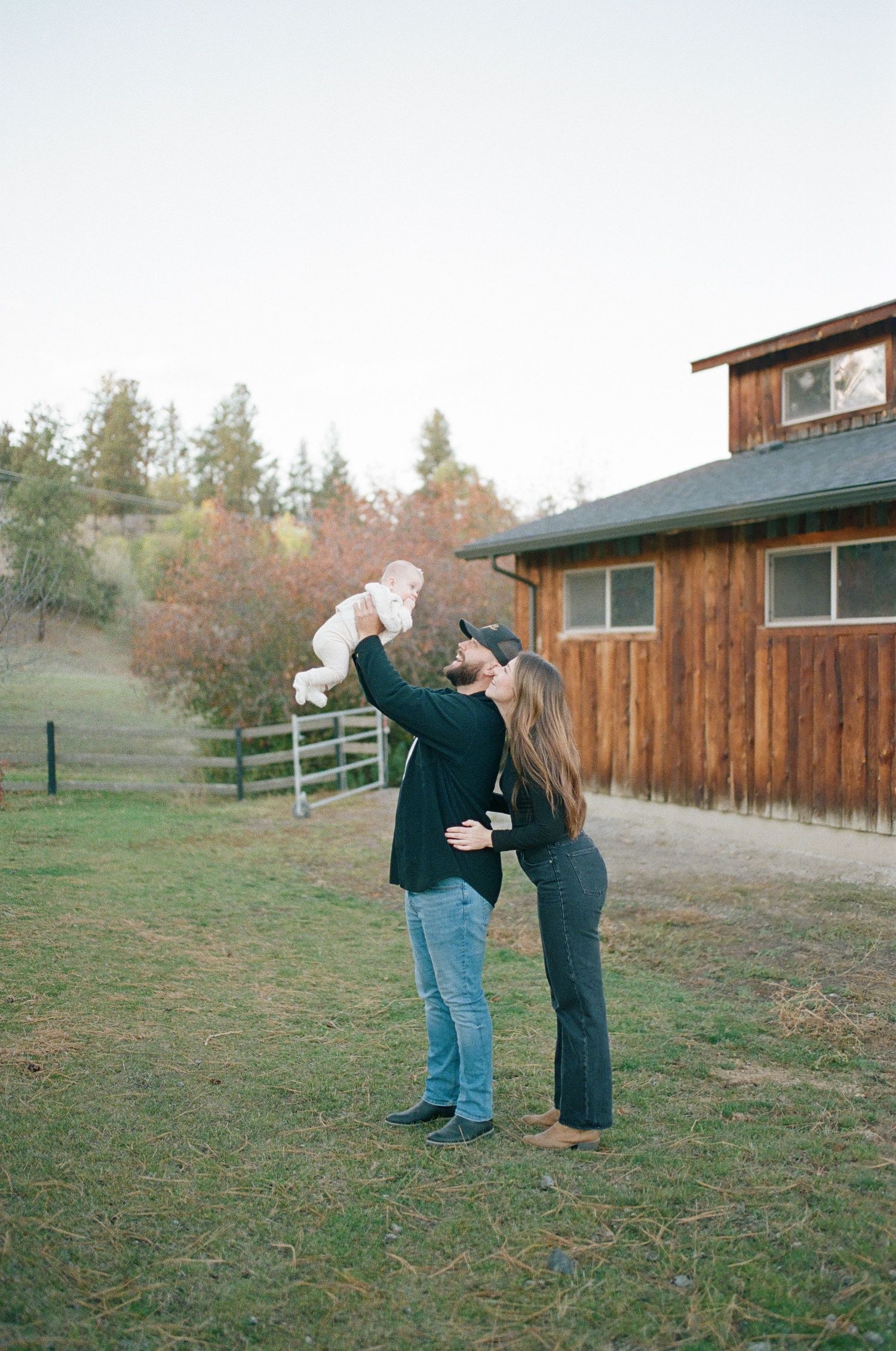 Two people hold a small white dog up in the air together near a rustic wooden barn in an autumn setting.