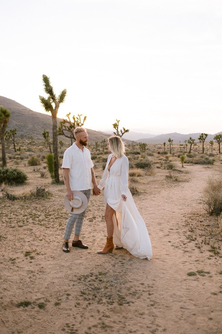 Two people in coordinated white outfits standing on desert path with mountains in background.