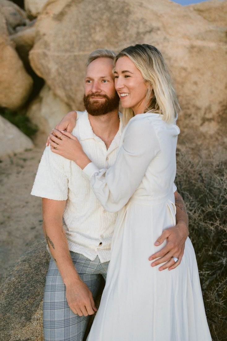 Couple in coordinated white outfits smiling and embracing by large desert rocks.
