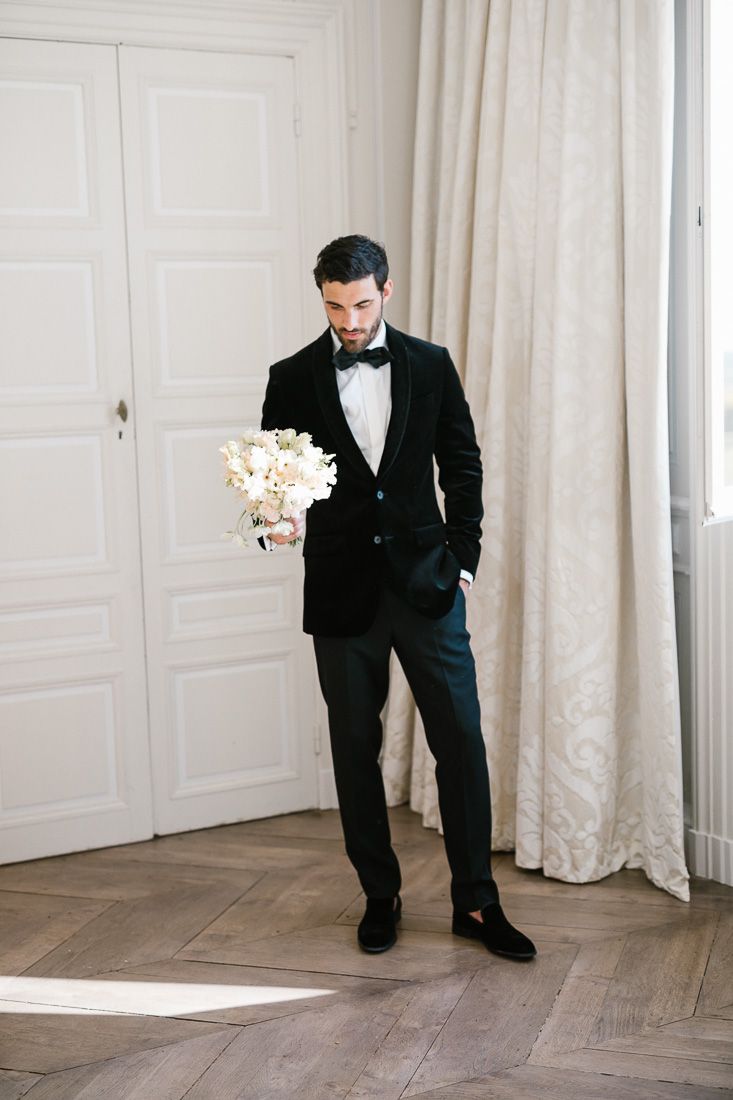 Groom in classic black tuxedo holds white bouquet while standing by white paneled wall.