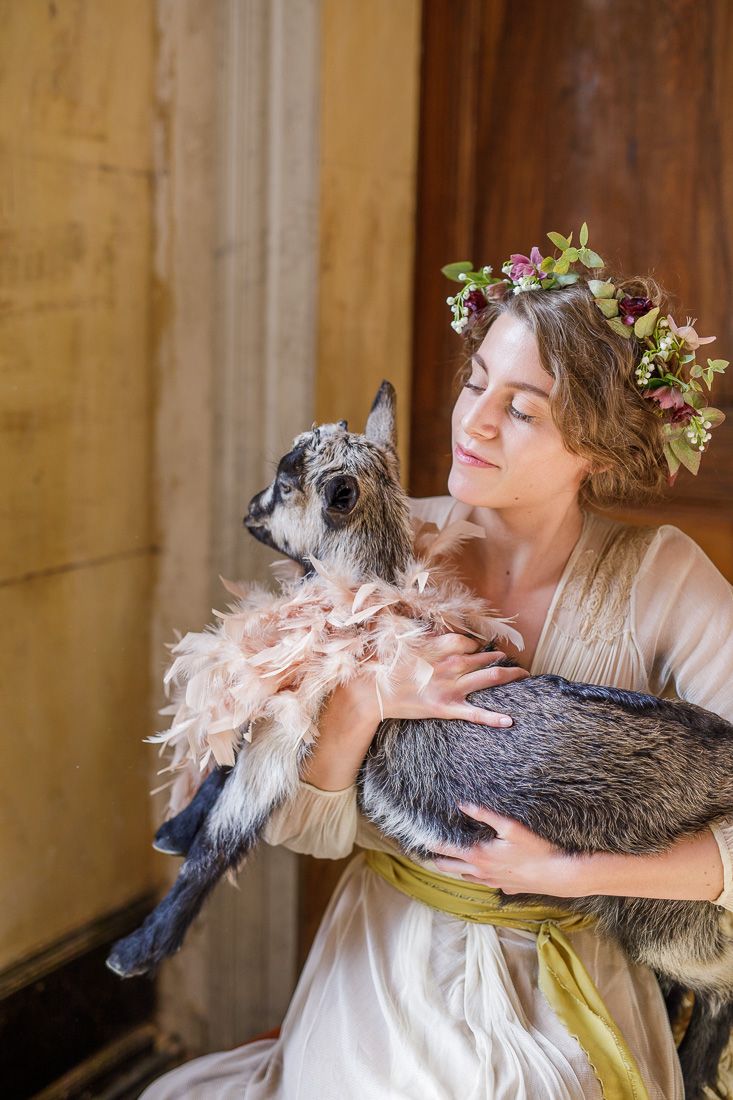Ethereal portrait of woman in flowing dress holding gray cat with floral crown.