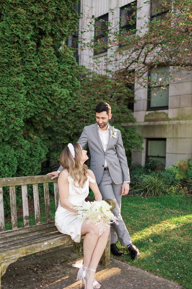 Romantic wedding photo on wooden bench against ivy-covered stone building.