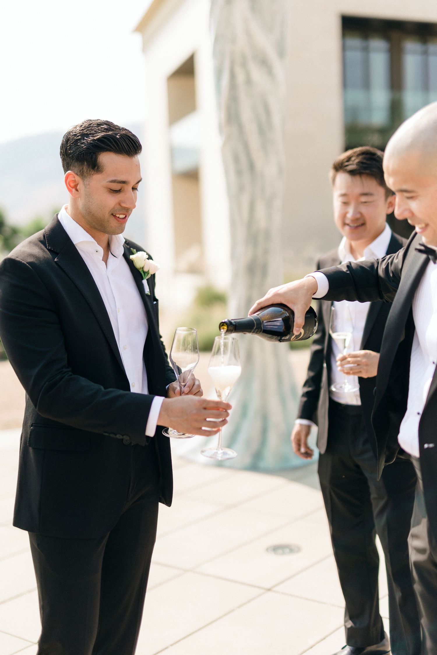 Groomsmen in black suits share champagne and laughter during wedding celebrations.