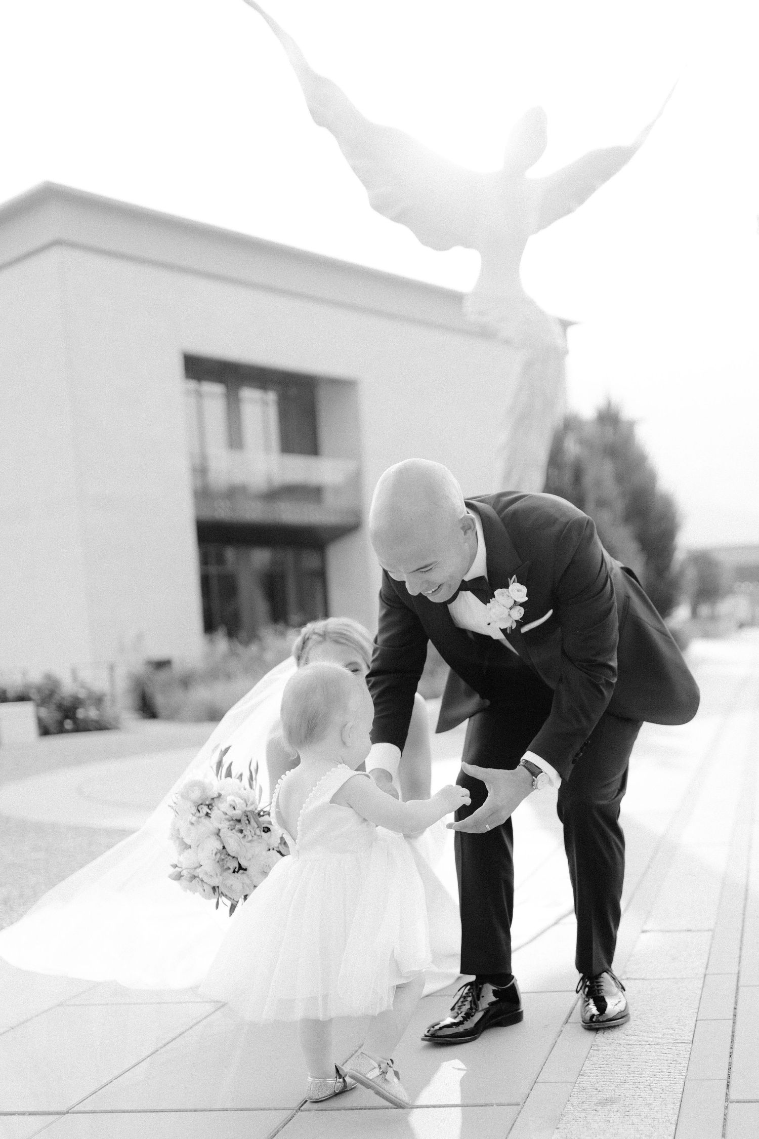 Sweet moment between a child in white and adult in formal wear near an angel statue.