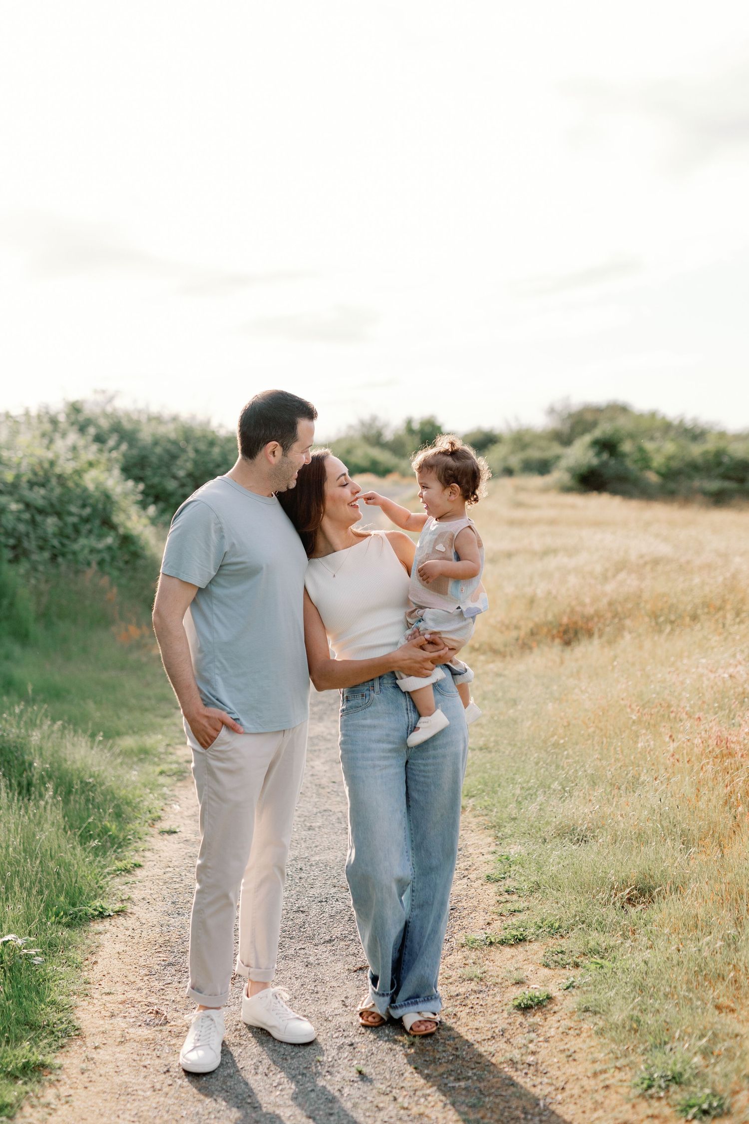 A family wearing casual outfits walks together along a dirt path through a sunlit meadow on a summer day.