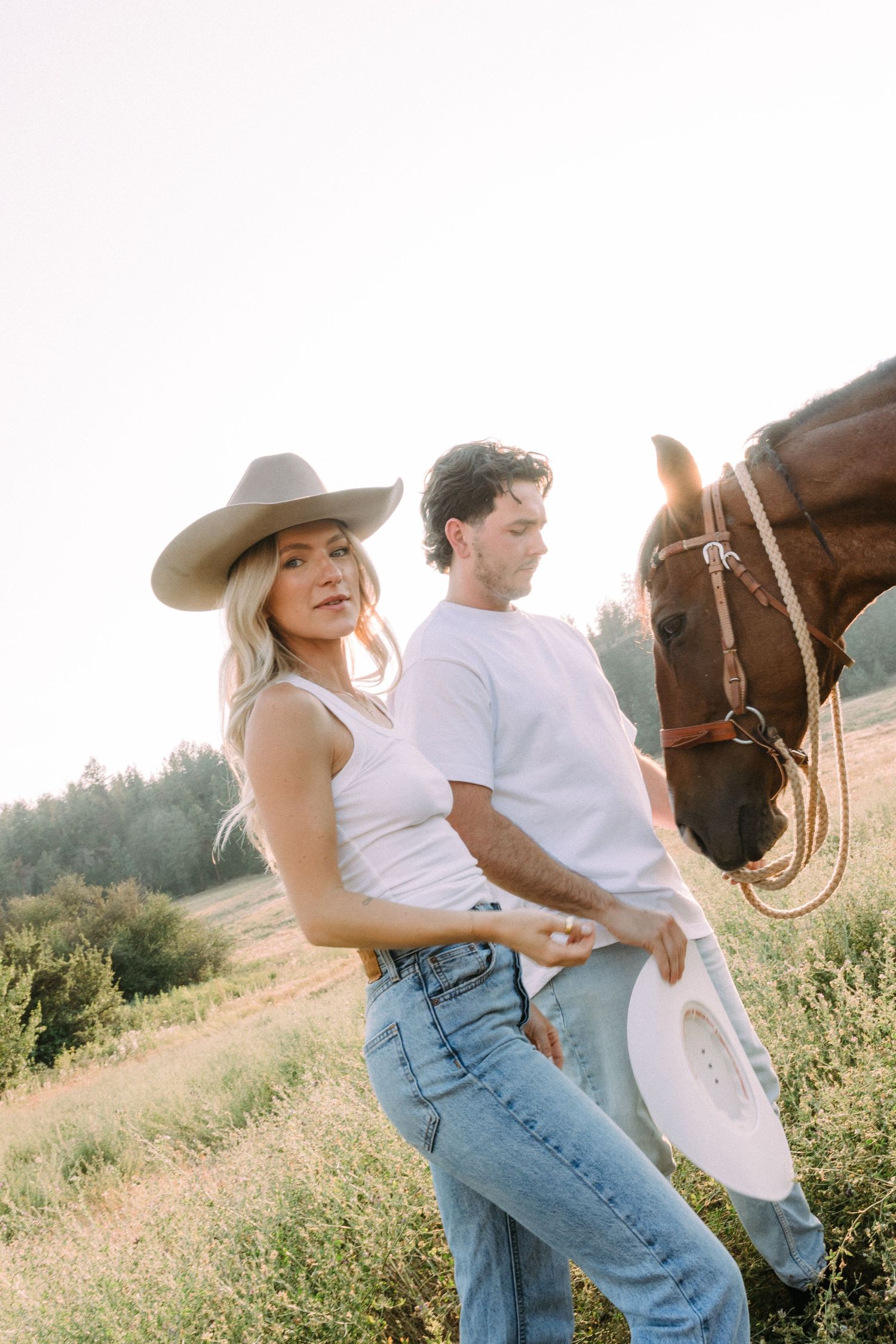 Couple in Western attire sharing a playful moment with their horse in a sunny field.