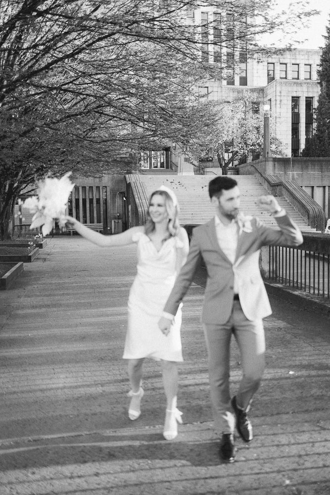 A joyful black and white photo of a couple jumping and celebrating on a walkway surrounded by trees.