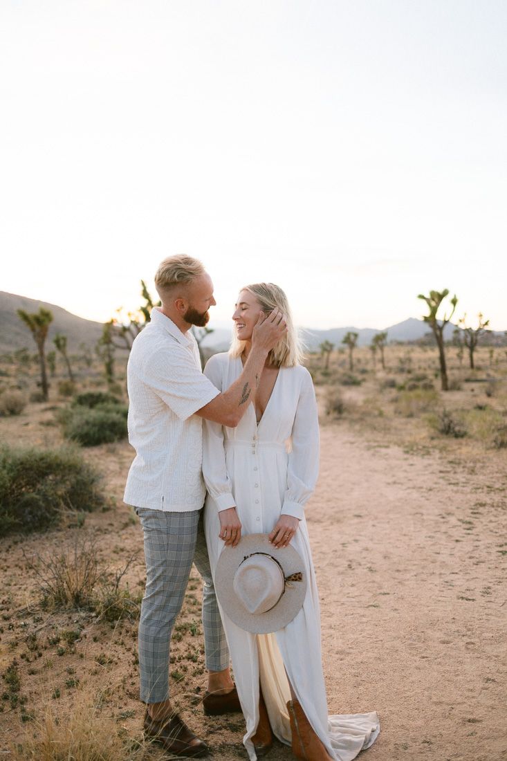 A couple in white outfits shares a tender moment on a desert path with Joshua trees at sunset.