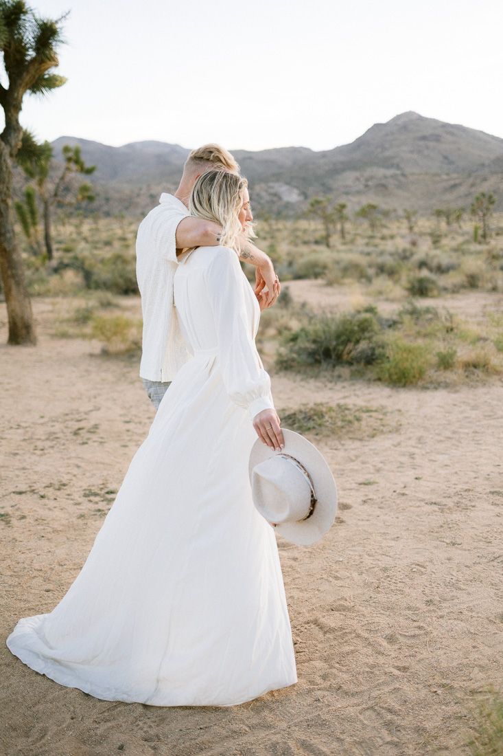 A flowing white dress catches the desert breeze as a couple stands among Joshua trees.