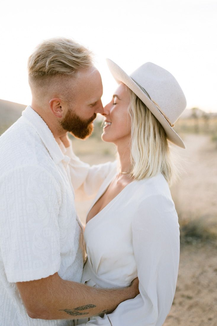 A romantic desert engagement photo of a couple in white outfits sharing an intimate moment at sunset.