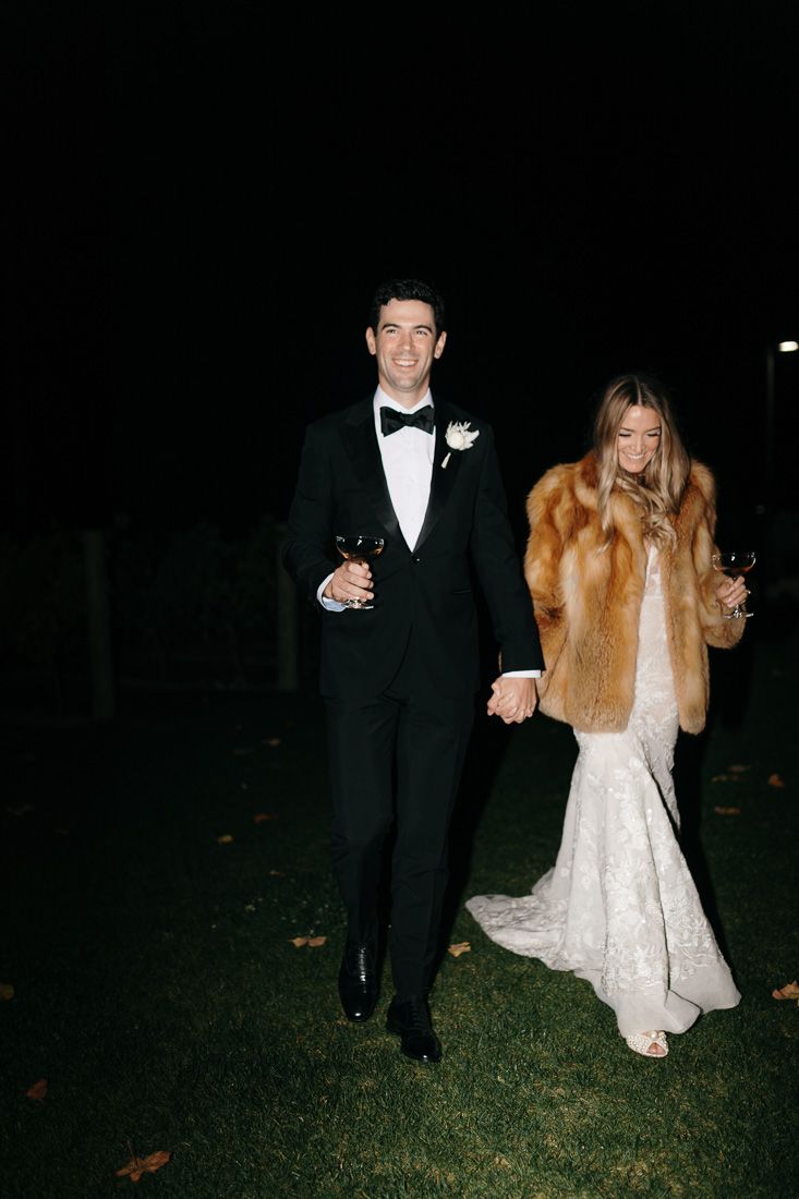 Newlyweds walking through a garden at night, bride in lace gown and fur coat.