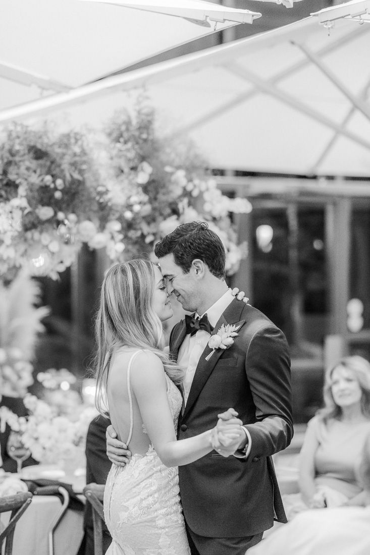 Newlyweds share their first dance under a white tent decorated with floral arrangements at an outdoor wedding reception.