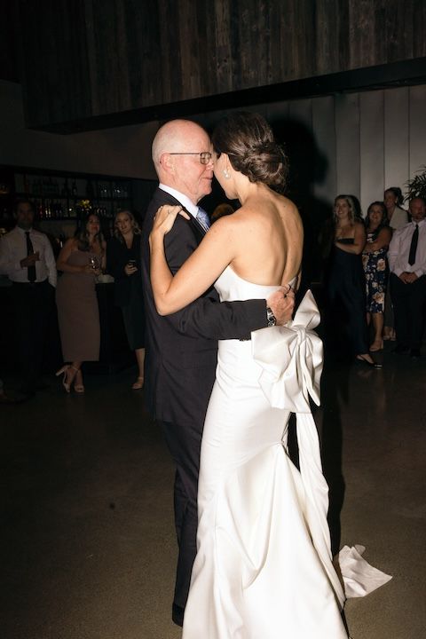 A couple shares a tender dance at their wedding reception, with the bride wearing a white gown with a bow detail.