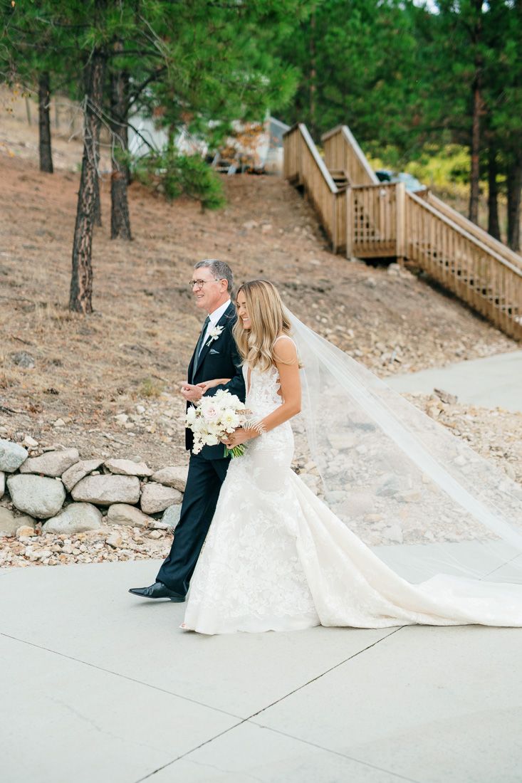A bride and her escort walk along a path with wooden stairs and pine trees in the background.