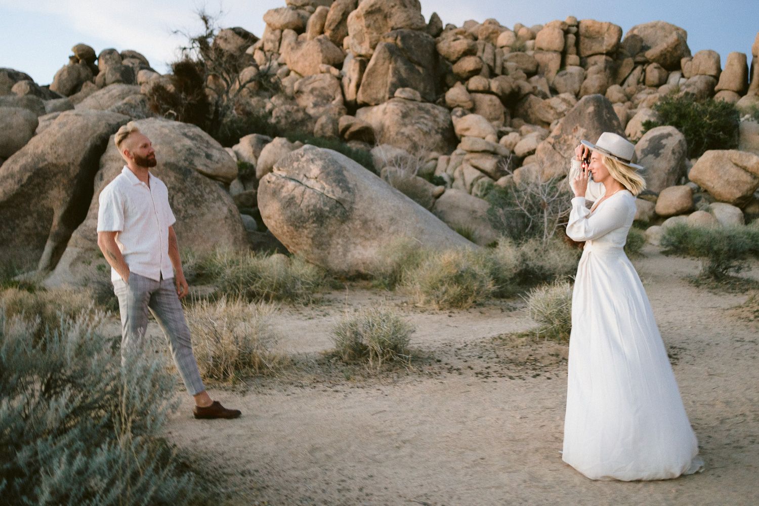 Couple in white outfits taking photos among large desert boulders during golden hour.
