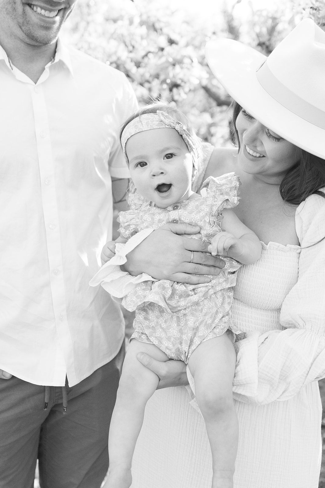 A black and white collage showing a joyful family moment with a baby in white outfits outdoors.
