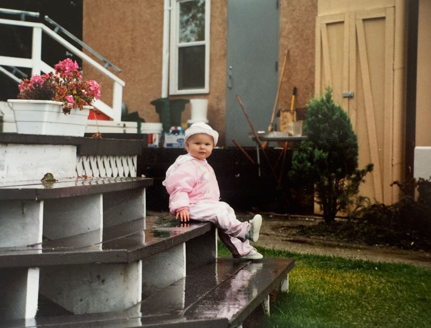 A small child in pink winter clothing sits on concrete steps with a wooden door and pink flowers visible in the background.