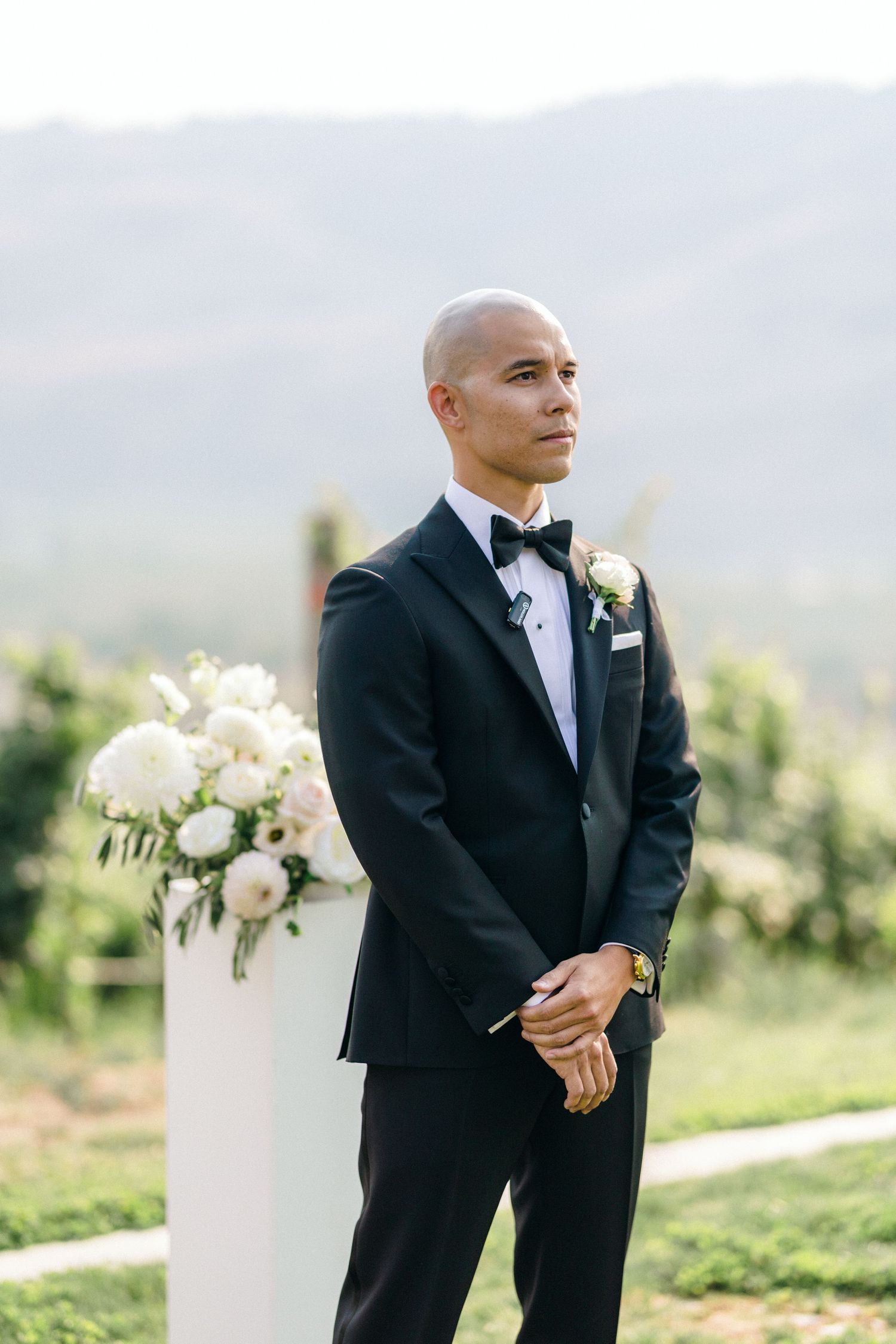 Groom in classic black tuxedo and bowtie stands in front of mountain landscape.