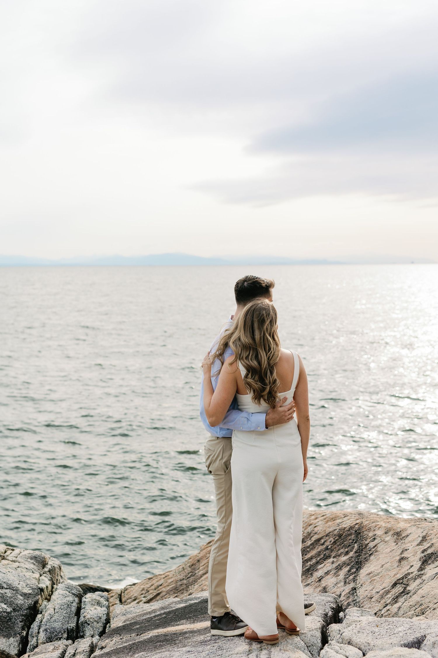 Two people embrace while standing on rocky coastal cliffs overlooking an ocean view at sunset.