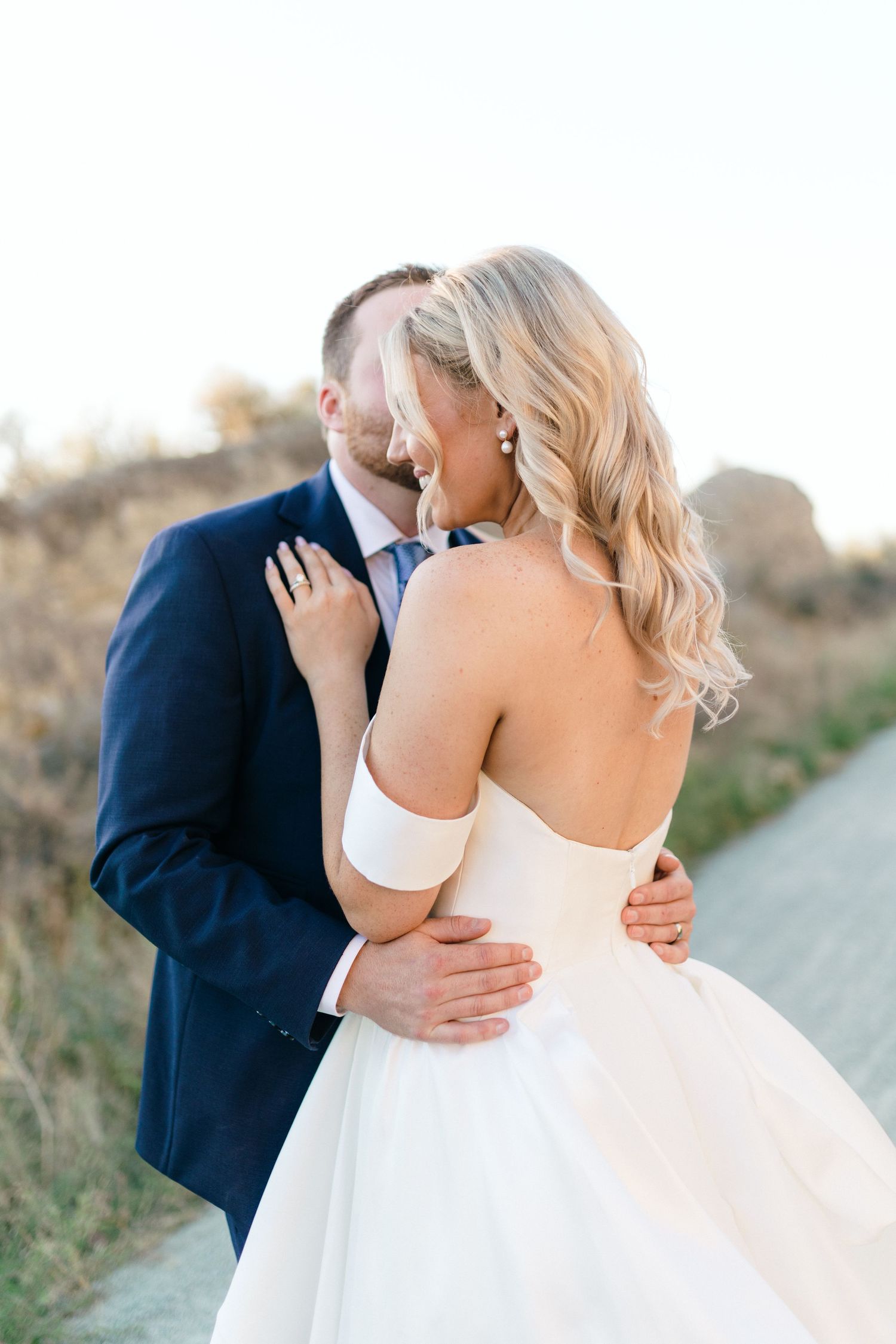 A romantic wedding couple embraces outdoors, with the bride in a white strapless gown and groom in navy suit.