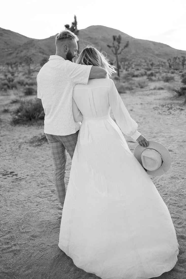 Black and white photo of a couple embracing while walking in a desert landscape.