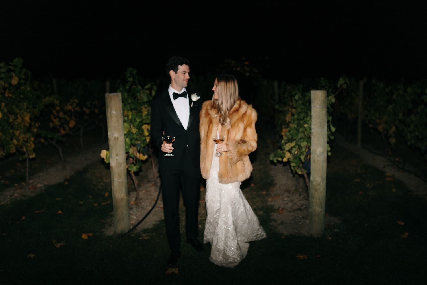 Elegantly dressed couple enjoying an evening moment between vineyard rows.