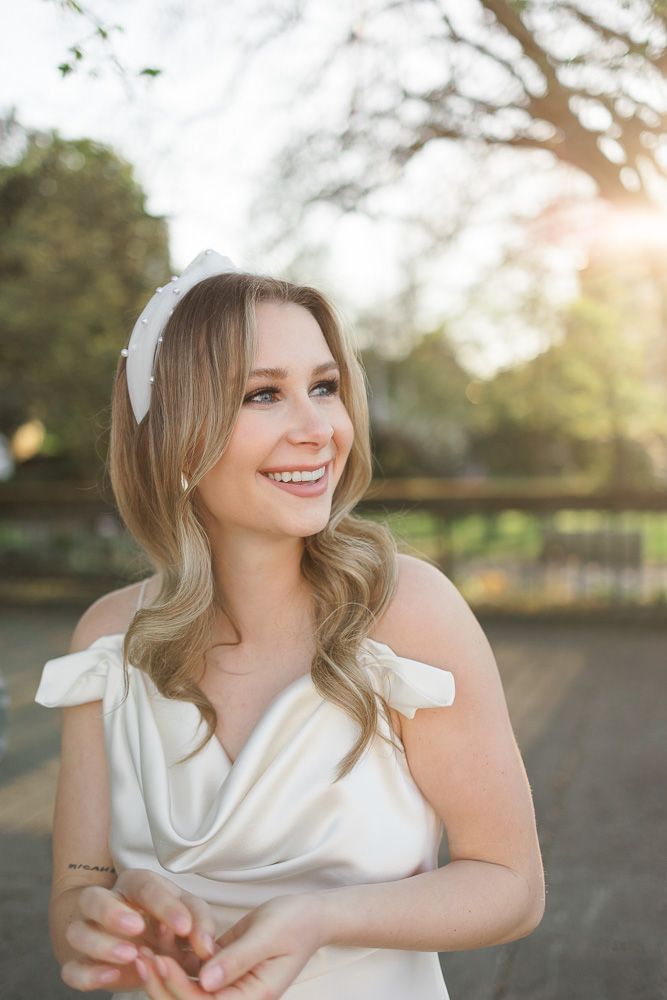 A bride in a white dress with bow details smiles while looking to the side in golden hour lighting.