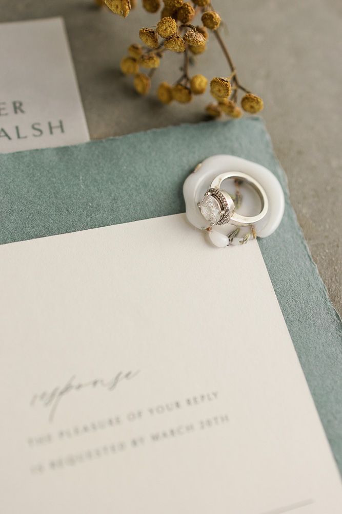 Wedding rings displayed on white stationery with sage green paper and golden dried flowers.