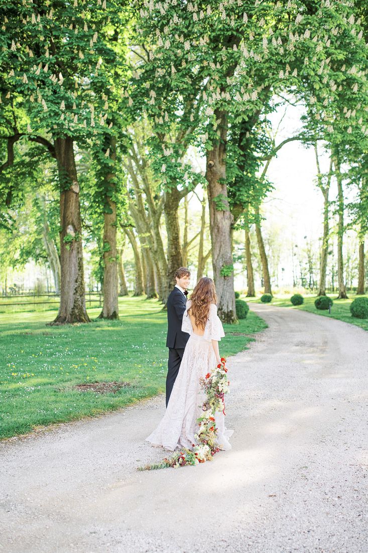 Romantic couple standing on a tree-lined path with trailing floral bouquet.