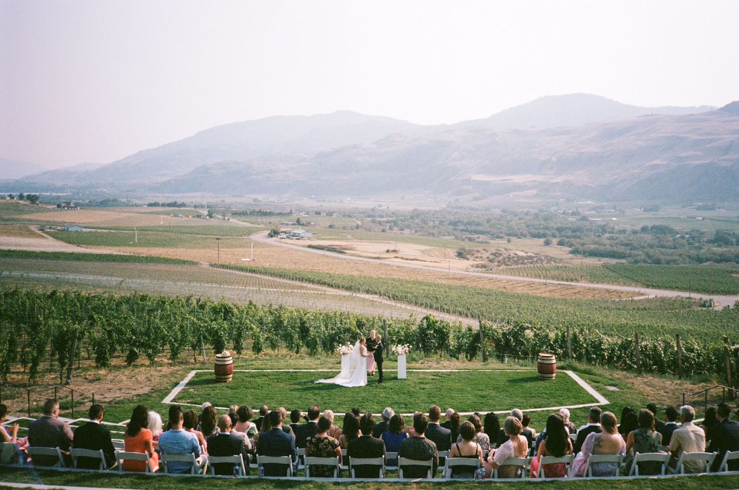 Panoramic view of an outdoor wedding ceremony with guests overlooking mountain vineyard landscape.