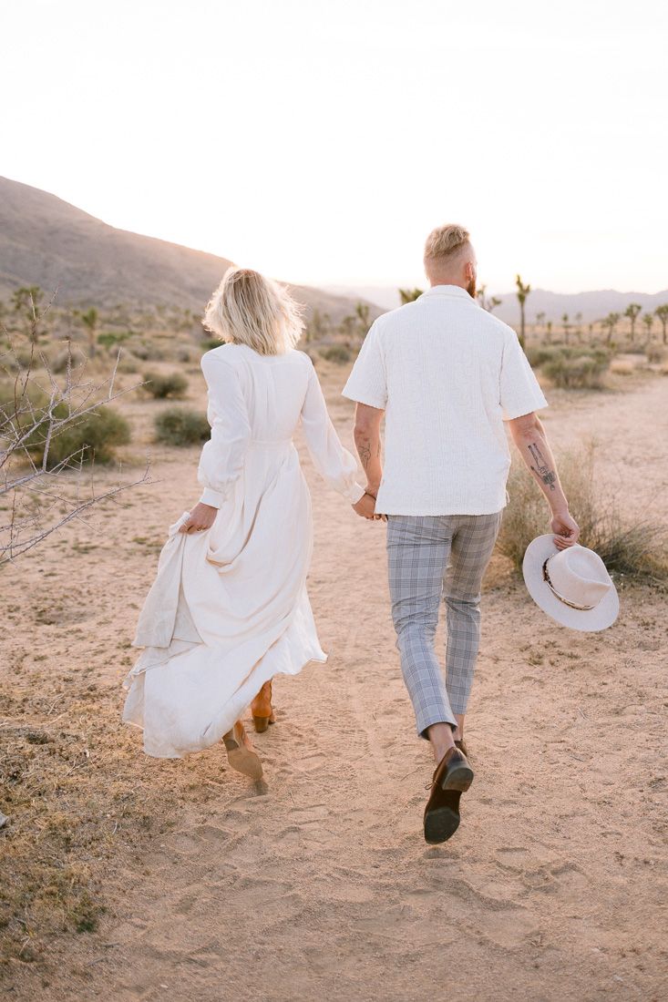 A couple walks hand in hand on a desert trail while holding a hat.