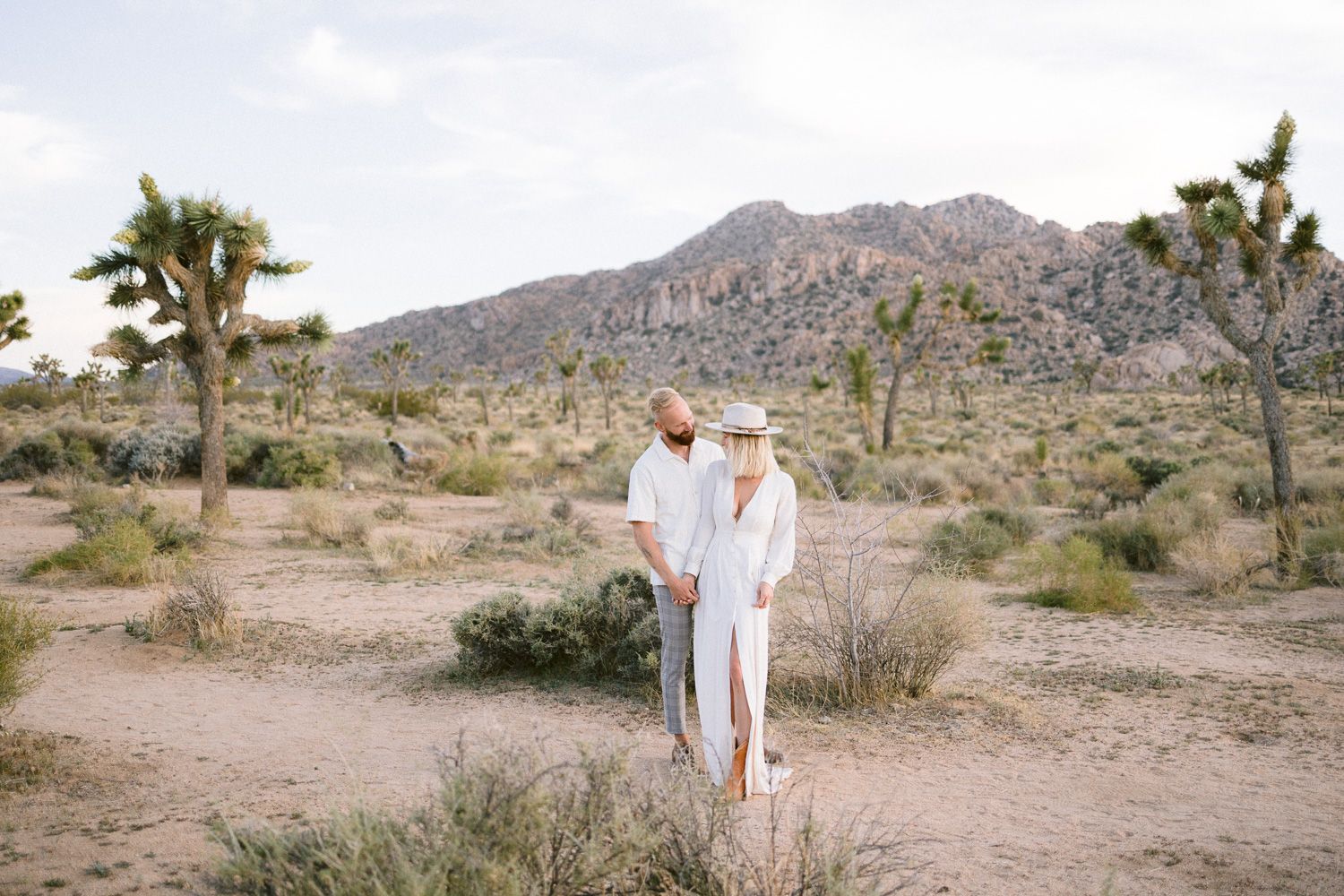 Wide angle desert portrait showing couple in white among Joshua trees and mountains.