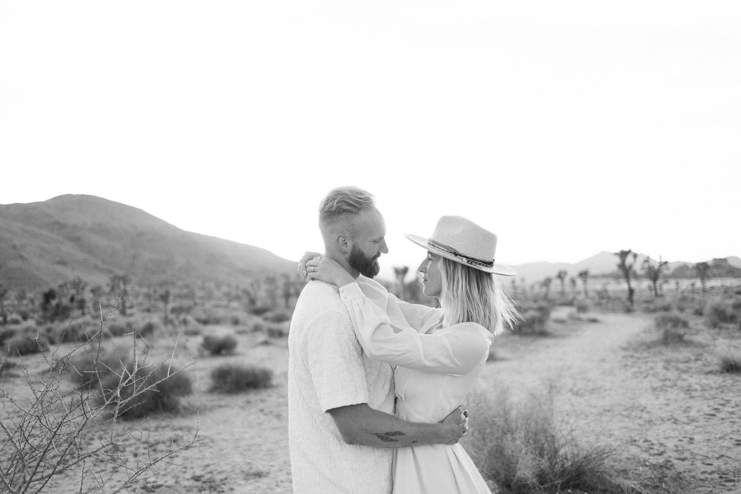 Black and white photo of a couple embracing in a desert landscape with mountains.