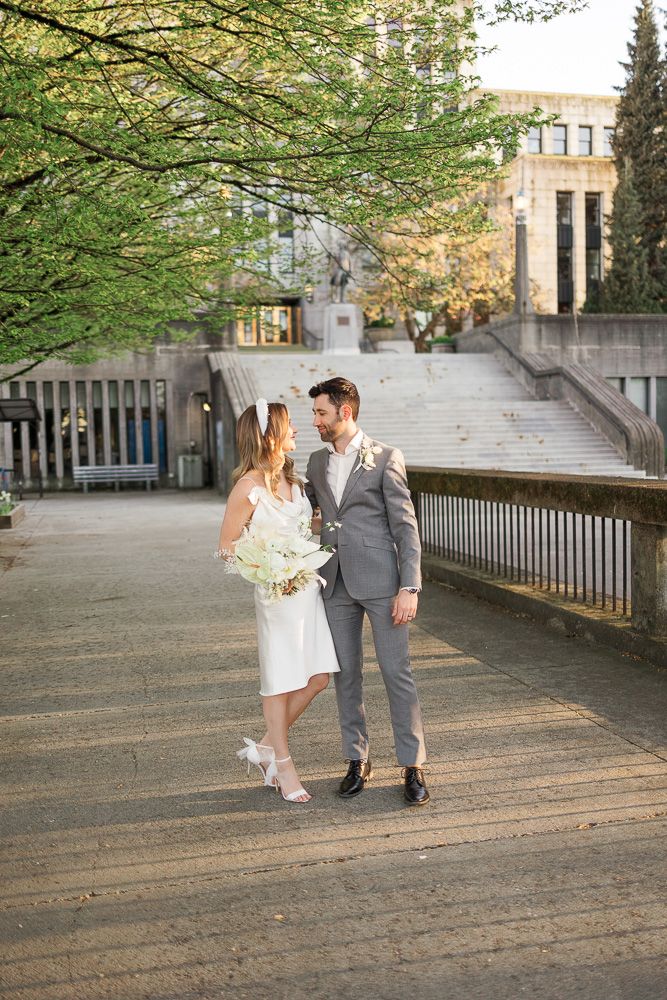 Newlyweds pose together on a concrete path with spring trees and architecture in the background.