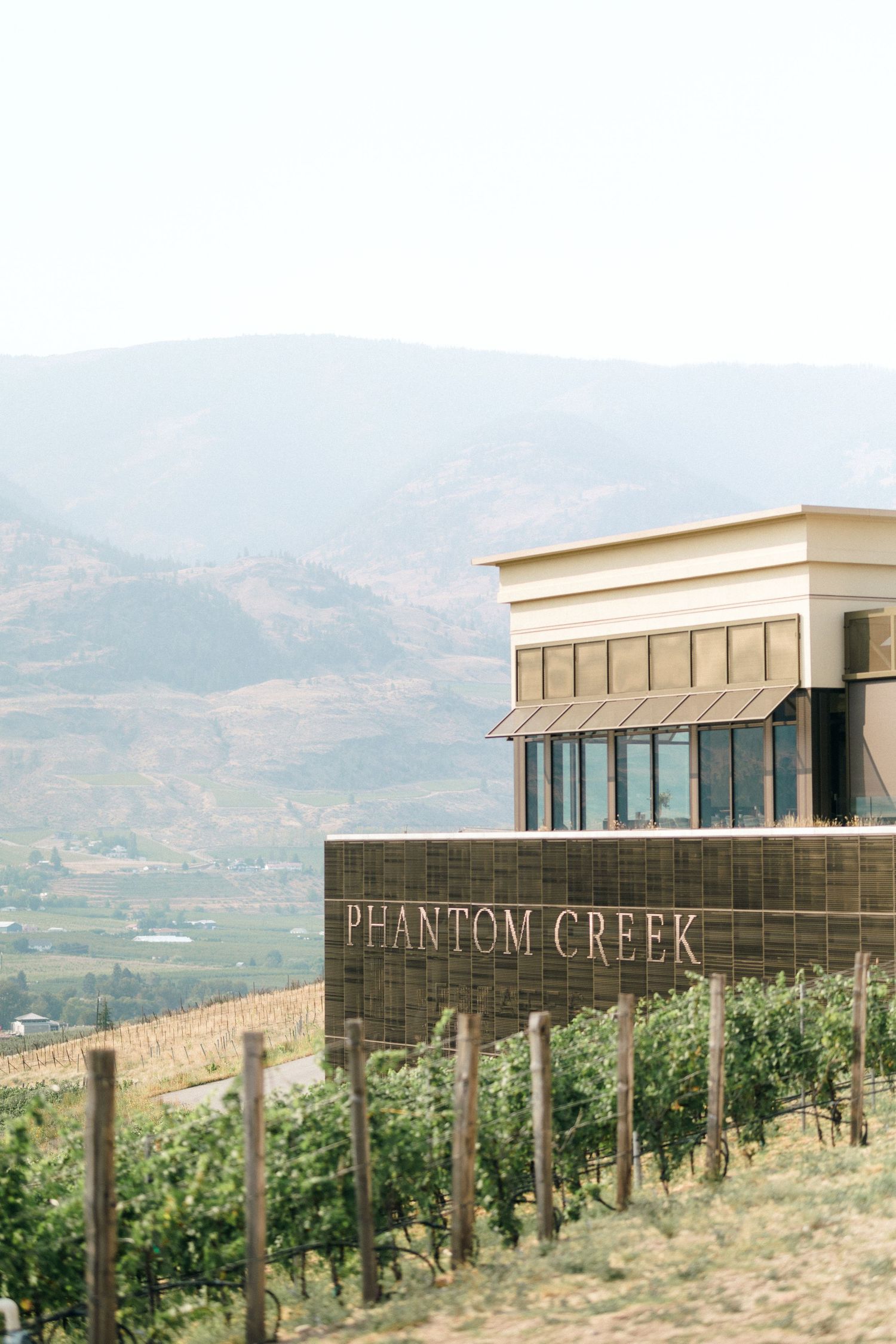 Modern winery building with Phantom Creek sign nestled among hillside vineyards with mountains in the background.