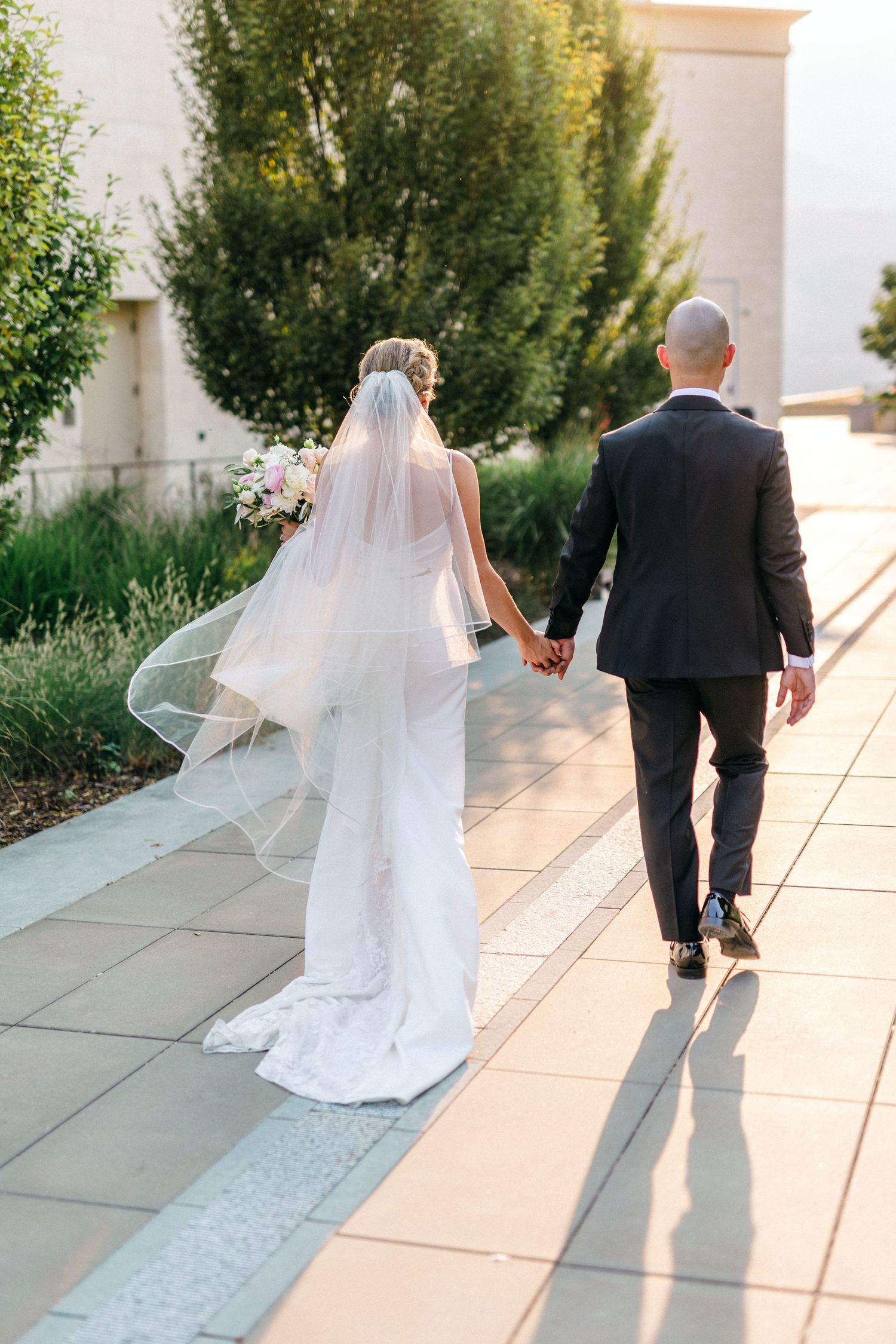 Couple walking along sunlit stone path with flowing wedding dress and veil.