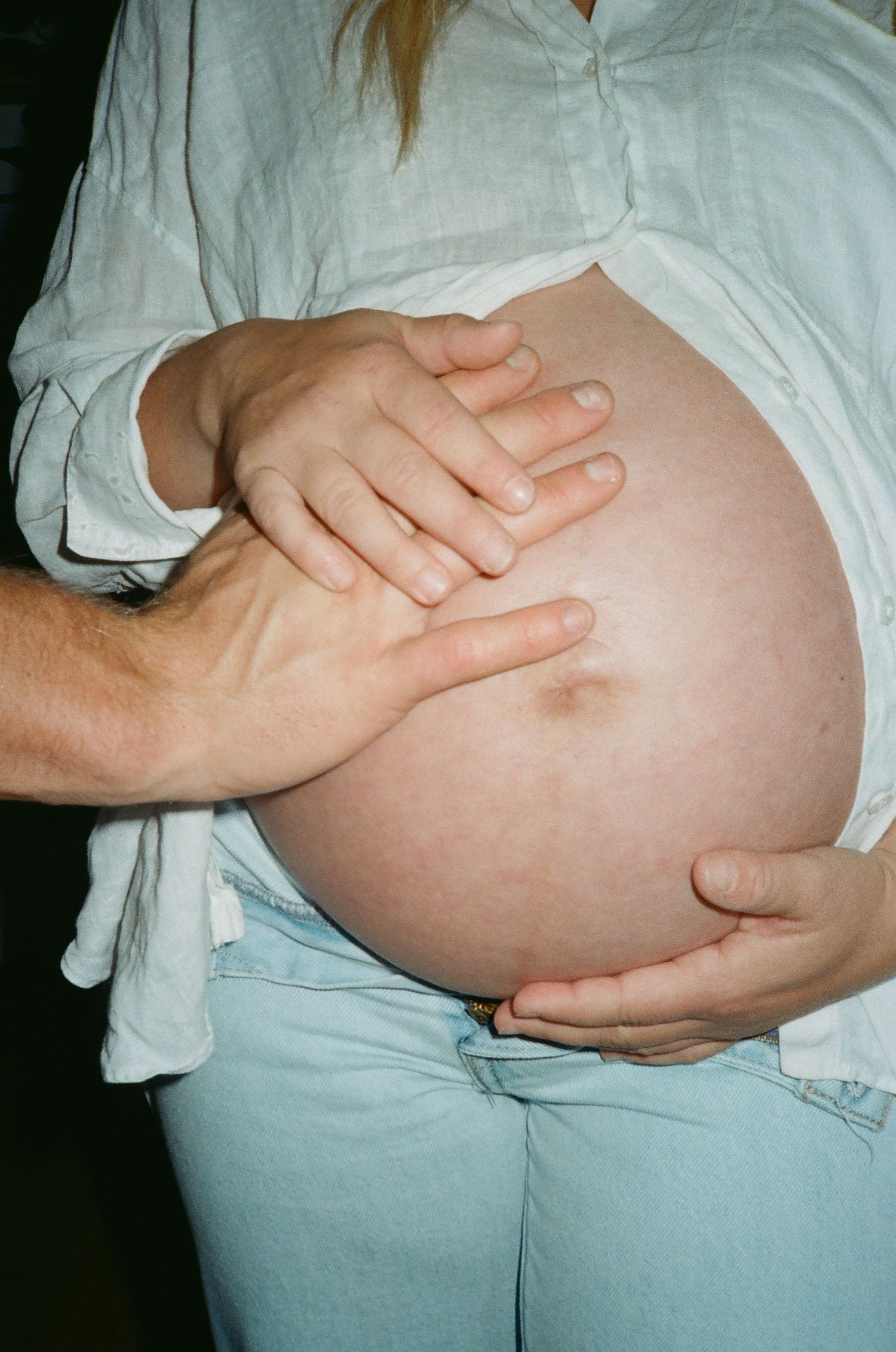 Hands tenderly cradle a pregnant belly against a white shirt and light jeans backdrop.
