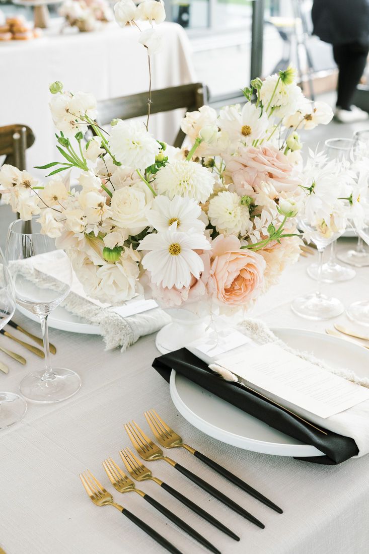 Elegant table setting with white and blush floral centerpiece, gold flatware, and black napkins on crisp white linens.