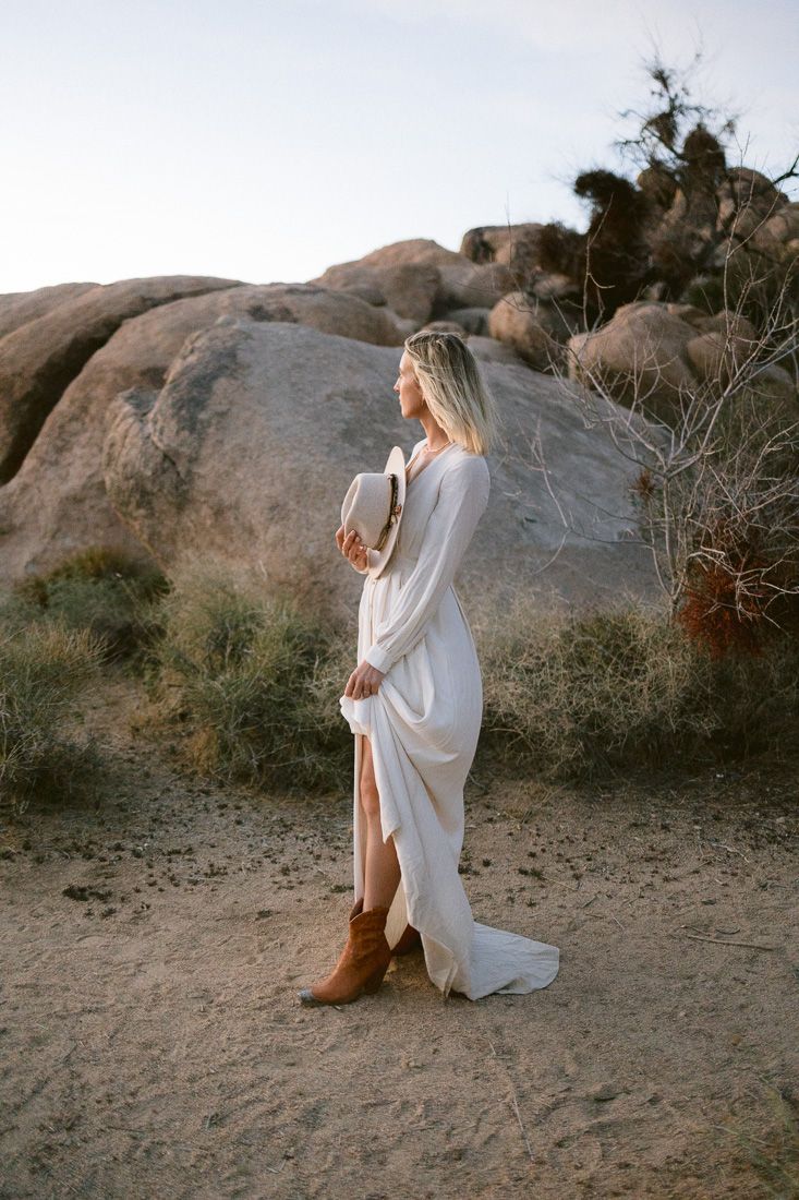 Person in flowing white dress and boots standing against desert rock formations at sunset.