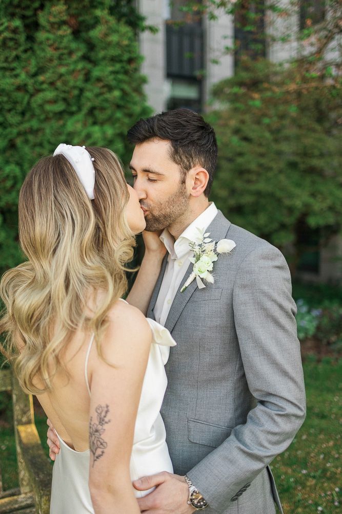 A tender wedding kiss moment captured in a garden setting with greenery and stone walls.