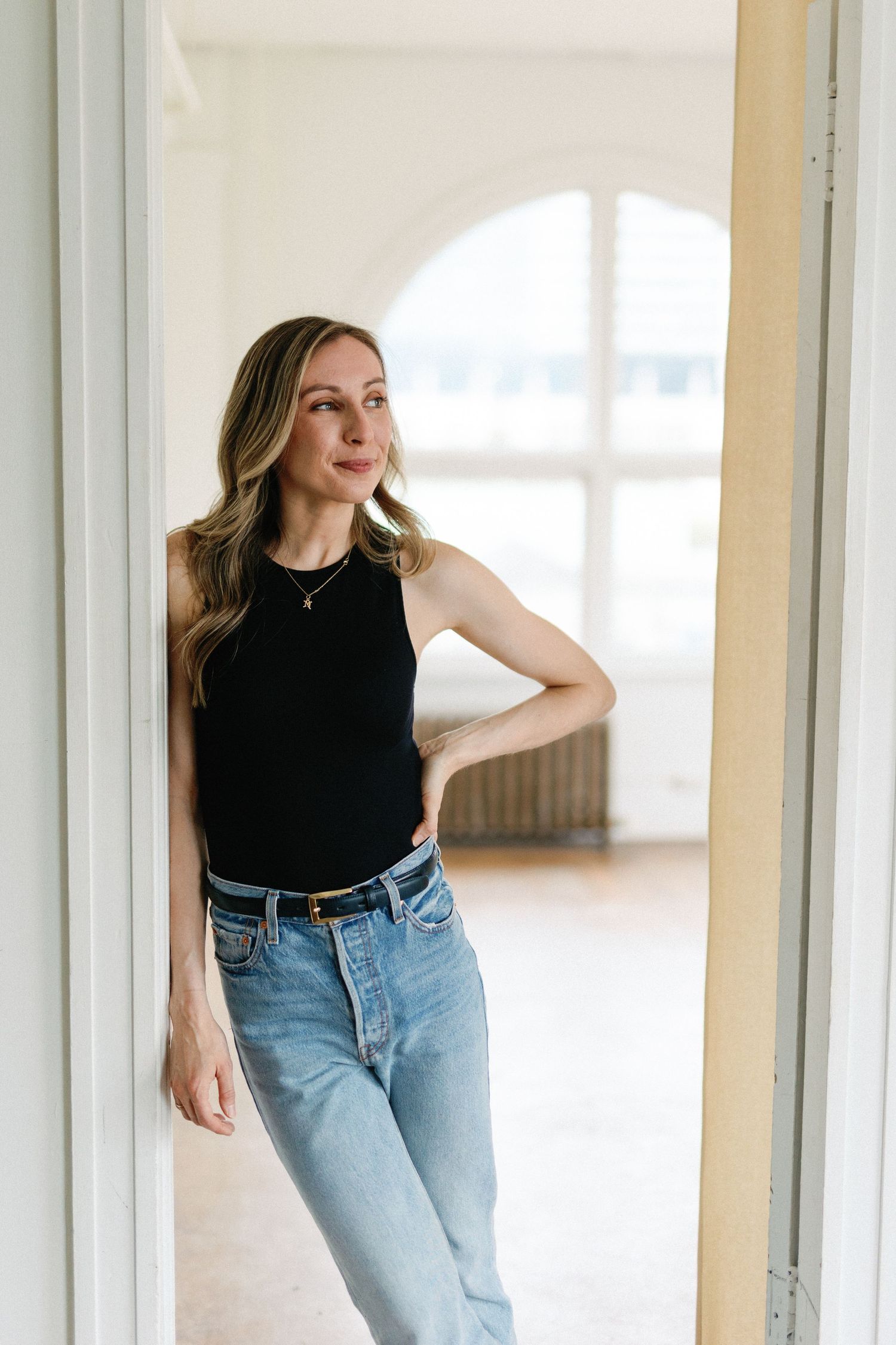 Woman in black sleeveless top and light wash jeans poses casually by white doorway in sunlit room.