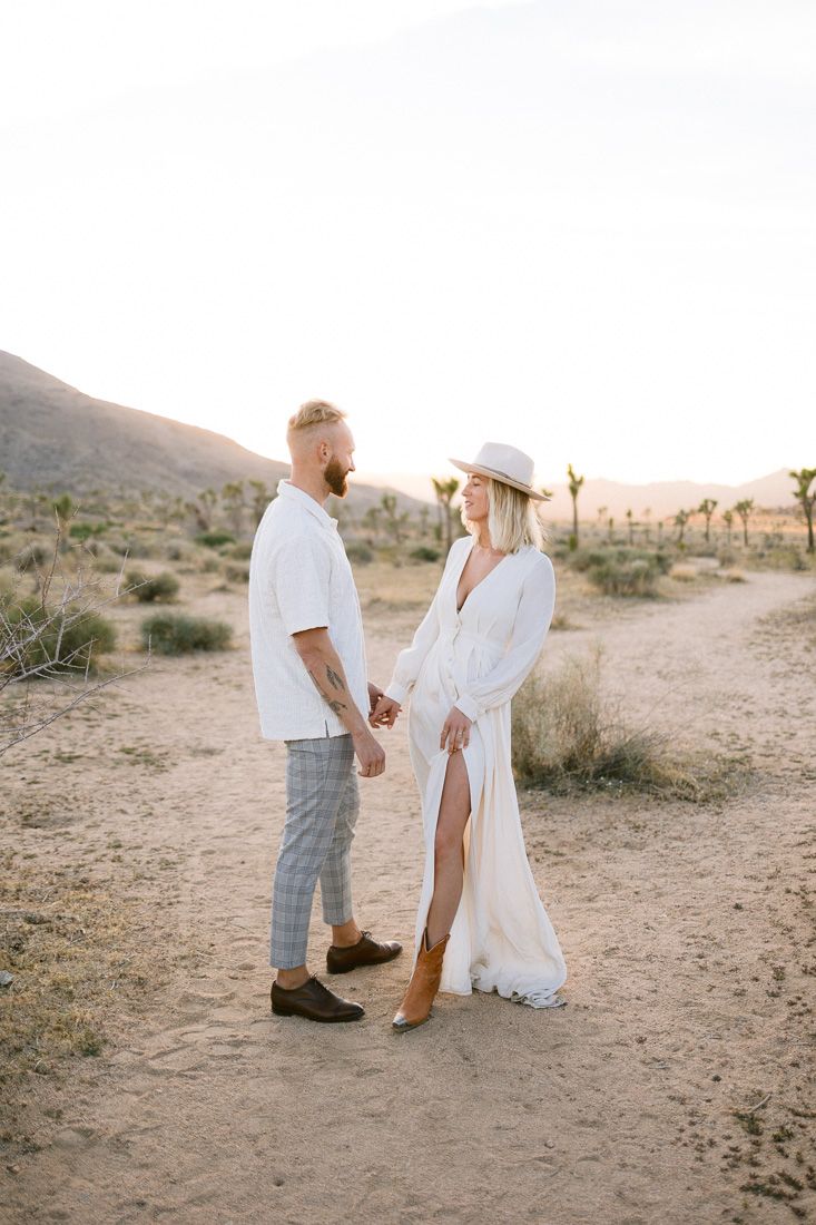 Sunset desert portrait of a couple in white outfits holding hands among Joshua trees.
