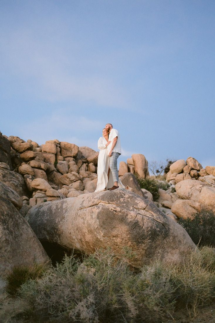 Couple in white standing atop large boulder formation against blue desert sky.