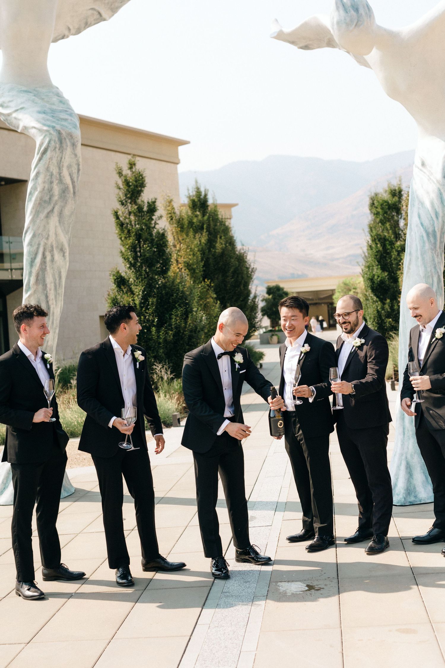 A wedding party in formal attire sharing laughs on an outdoor terrace with mountain views.