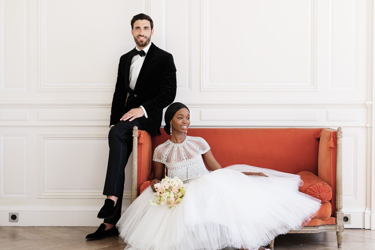 Elegant wedding couple poses on orange vintage sofa in white room with bride in tea-length tulle dress.