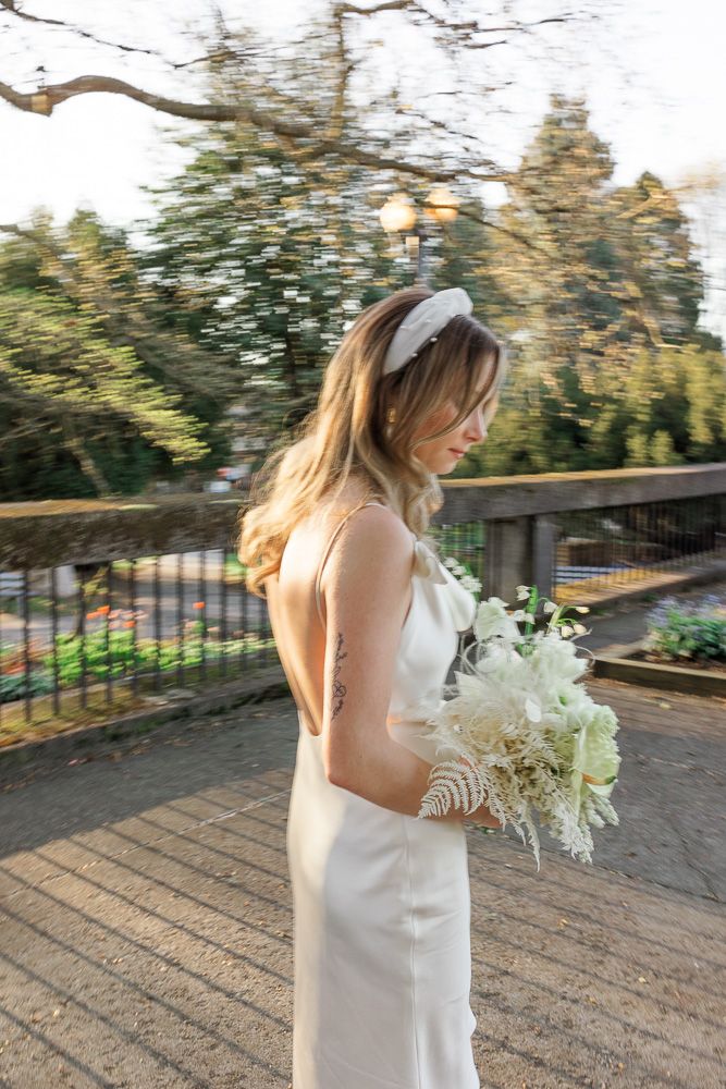 Bride in elegant white dress holds bouquet while standing in outdoor garden setting.