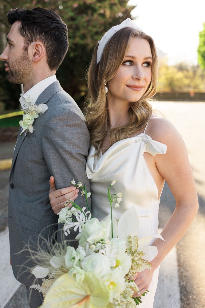 Elegant wedding couple in grey suit and white dress with spring bouquet outdoors.