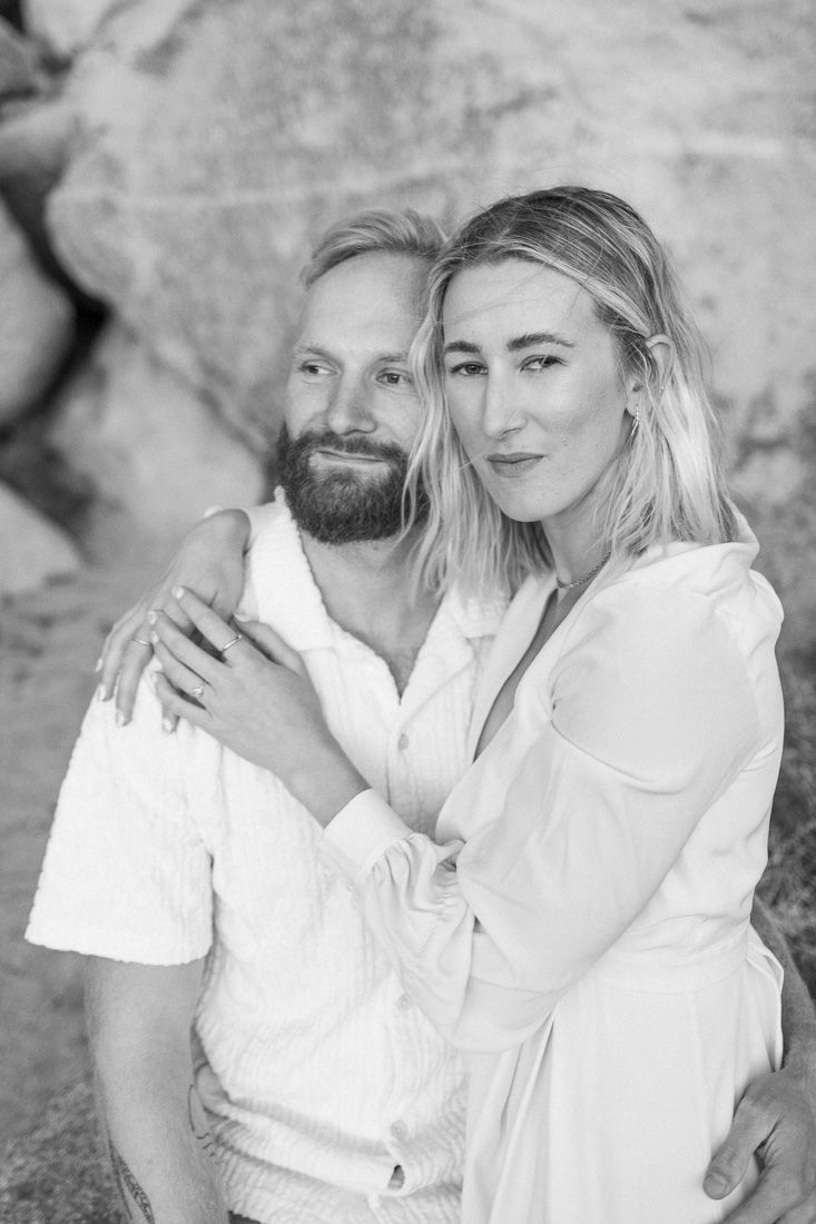 Black and white portrait of a couple in white clothing embracing against a rocky backdrop.