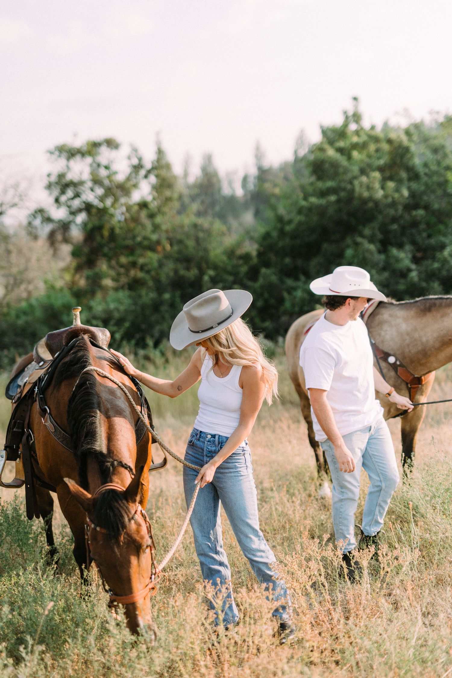 A couple spends time with their horses in a sun-drenched meadow wearing casual western attire.